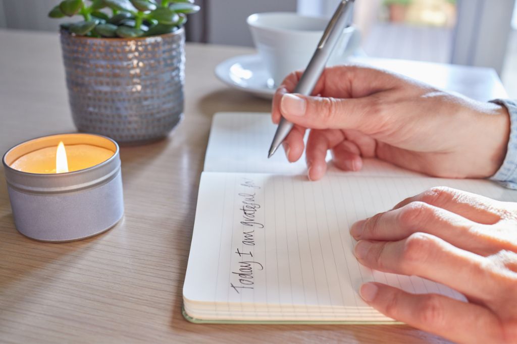 A person writes in a gratitude journal at a desk holding a candle. Journaling can also help identify feelings of expat homesickness.