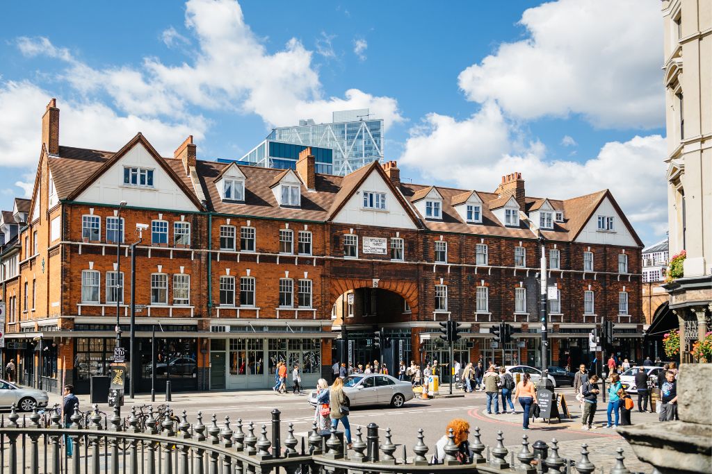 Exterior of Old Spitalfields Market in London. Pedestrians cross the road around the market building. The market is covered, making it an ideal spot to visit on a rainy day in London.
