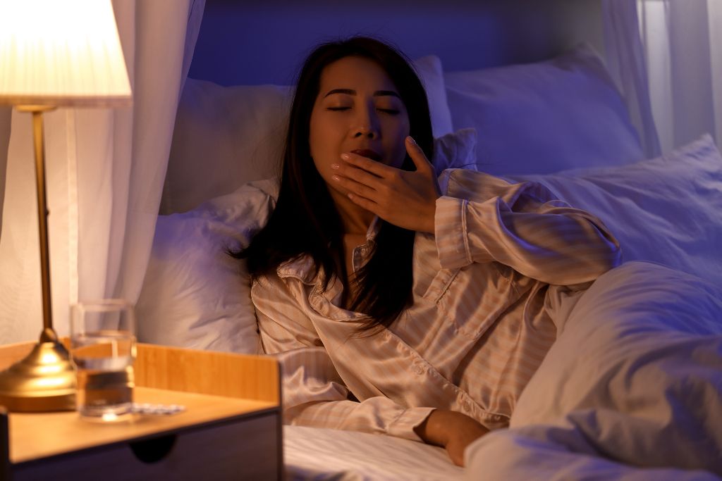 Woman yawning in bed with melatonin and a glass of water next to her on the nightstand.